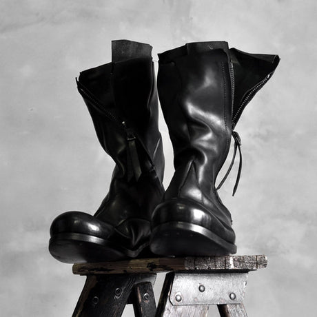 Pair of black high shaft deconstructed leather boots displayed on stool showing zipper and sculptural silhouette, Alternative Darkwear